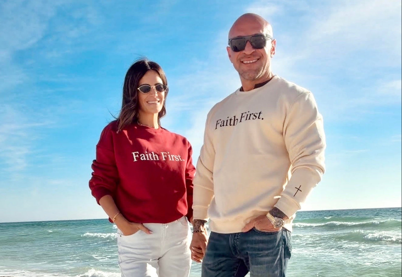 Two people wearing 'Faith First' sweatshirts standing on a beach with blue sky and ocean.